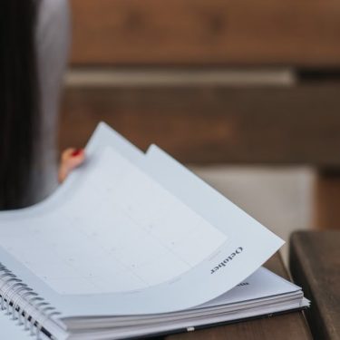 Crop unrecognizable female student with dark hair in white blouse turning pages of notepad at wooden table in park