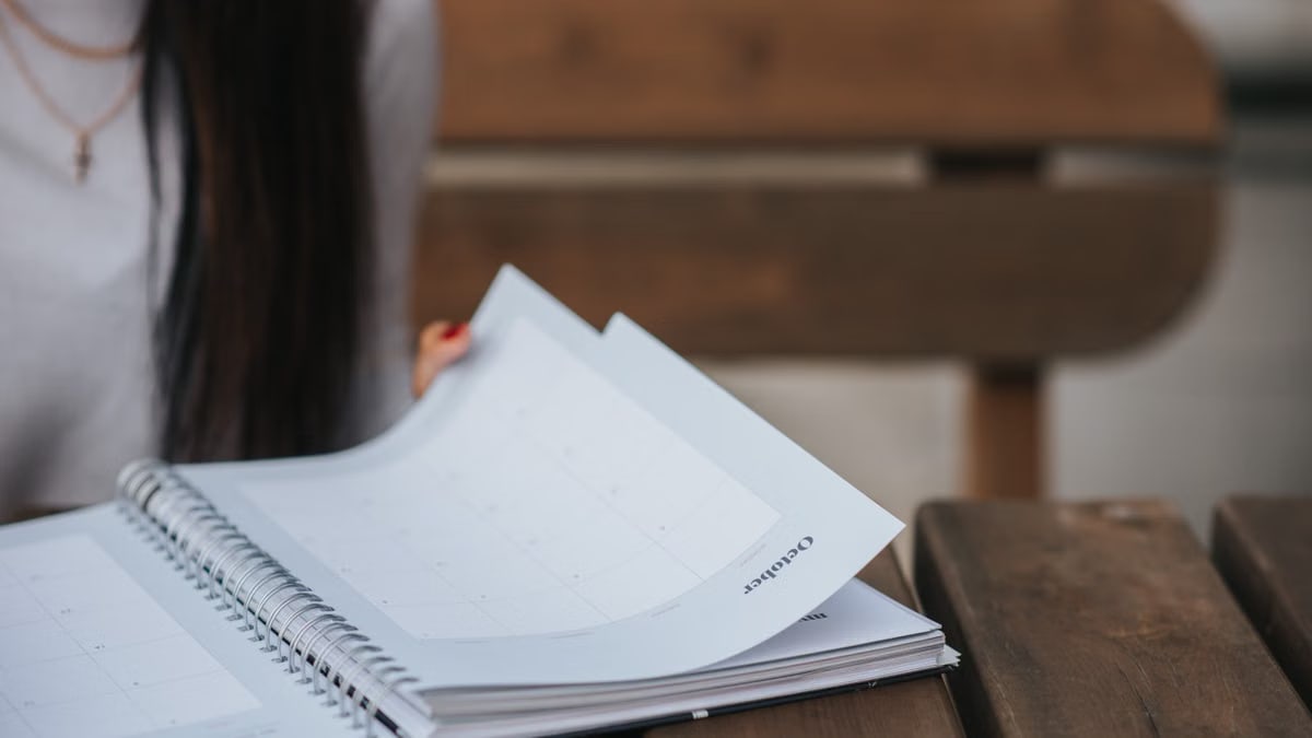 Crop unrecognizable female student with dark hair in white blouse turning pages of notepad at wooden table in park